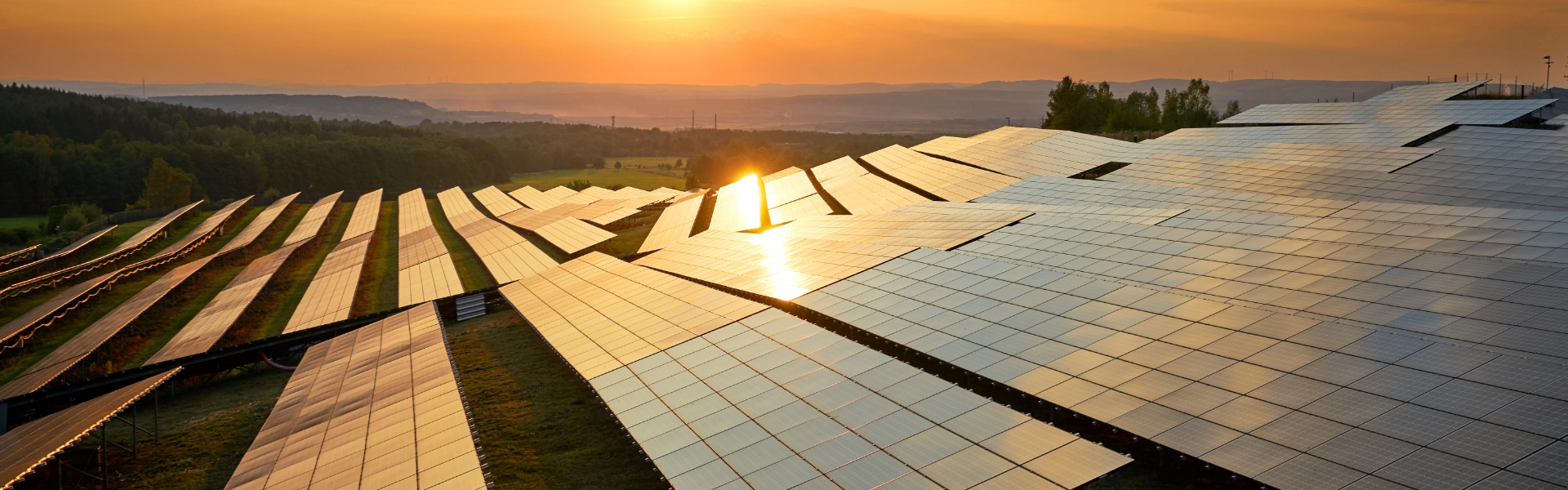 Rows of solar panels with green mountains and sun setting in the background